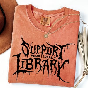 May include: A terracotta-colored short-sleeve t-shirt featuring the black text "SUPPORT YOUR LOCAL LIBRARY" in a stylized, jagged font. The shirt has a crew neckline and a Comfort Colors brand label visible at the inner collar.