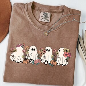May include: A brown Comfort Colors t-shirt featuring a graphic of four cartoon ghosts with floral arrangements and pumpkins. A silver necklace with a pendant is draped across the shirt.