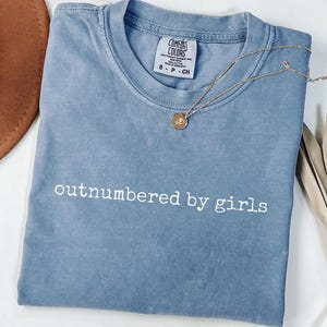 May include: A blue t-shirt with the phrase "outnumbered by girls" in white lettering. A gold necklace with a pendant rests on the shirt. The shirt is folded and placed on a white surface.