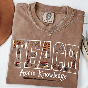 May include: A brown short-sleeved t-shirt with a crew neckline. The front features the word TEACH in large white block letters filled with magical illustrations including books, wands, and owls. Below, the text reads Accio Knowledge.