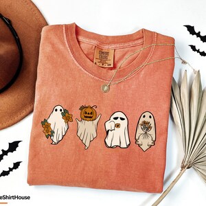 May include: A peach-coloured t-shirt with a graphic of three ghosts. The ghost on the left is holding flowers, the middle ghost is holding a pumpkin, and the ghost on the right is holding a bouquet of flowers. The text "COMFORT COLORS" is on the tag.