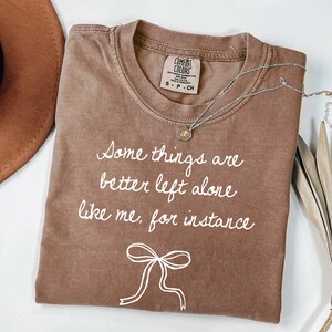 May include: A brown Comfort Colors t-shirt with the text "Some things are better left alone like me, for instance" in white script. A silver necklace with a pendant is draped across the shirt. A brown hat is visible in the background.