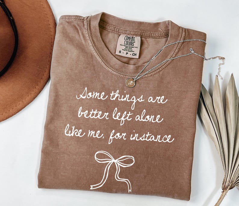 May include: A brown Comfort Colors t-shirt with the text "Some things are better left alone like me, for instance" in white script. A silver necklace with a pendant is draped across the shirt. A brown hat is visible in the background.