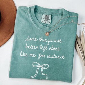 May include: A teal Comfort Colors t-shirt with the text "Some things are better left alone like me, for instance" in white script. A gold necklace with a pendant and a white bow graphic are also visible.