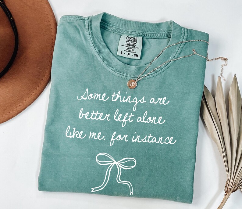May include: A teal Comfort Colors t-shirt with the text "Some things are better left alone like me, for instance" in white script. A gold necklace with a pendant and a white bow graphic are also visible.