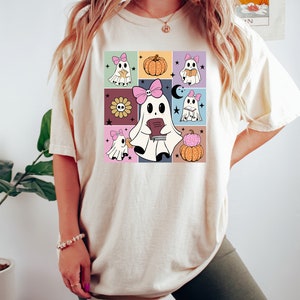 May include: Cream-colored t-shirt featuring a colorful grid design with cartoon ghosts, pumpkins, and flowers. The ghosts wear pink bows and hold books. The shirt is a casual, comfortable style.