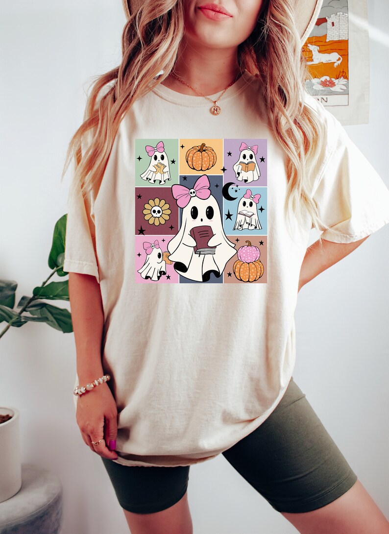May include: Cream-colored t-shirt featuring a colorful grid design with cartoon ghosts, pumpkins, and flowers. The ghosts wear pink bows and hold books. The shirt is a casual, comfortable style.