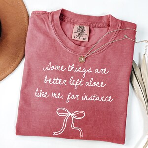 May include: A red-toned t-shirt with the text "Some things are better left alone like me, for instance" in white script. A gold necklace with a pendant rests on the shirt. A brown hat and dried leaves are in the background.