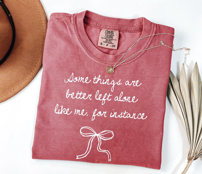 May include: A red-toned t-shirt with the text "Some things are better left alone like me, for instance" in white script. A gold necklace with a pendant rests on the shirt. A brown hat and dried leaves are in the background.