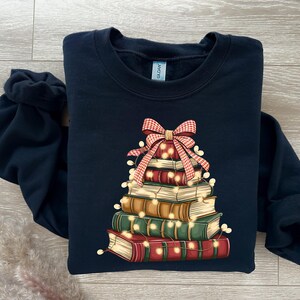 May include: A navy blue sweatshirt features a stack of books decorated as a Christmas tree. A red and white plaid bow sits atop the books, with string lights wrapped around them. The books are various colors.
