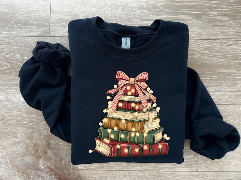 May include: A navy blue sweatshirt features a stack of books decorated as a Christmas tree. A red and white plaid bow sits atop the books, with string lights wrapped around them. The books are various colors.