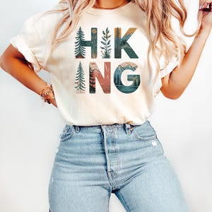 May include: A white t-shirt with the word "HIKING" printed on it in colorful letters. Each letter is filled with a nature scene, including trees, mountains, and a lake.
