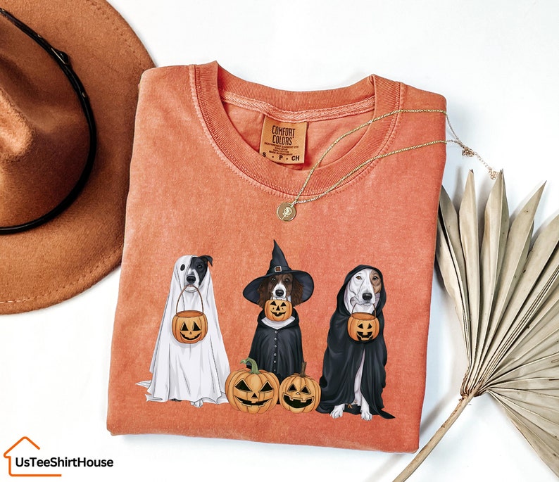 May include: A peach-coloured t-shirt featuring a graphic of three dogs dressed up for Halloween. The dogs are holding jack-o'-lanterns and are wearing costumes. The text "COMFORT COLORS" is on the tag.