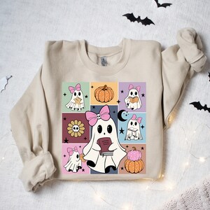 May include: Beige sweatshirt featuring a colorful graphic of cartoon ghosts with pink bows, pumpkins, and flowers. The design is set against a grid of squares with a retro aesthetic, perfect for Halloween.