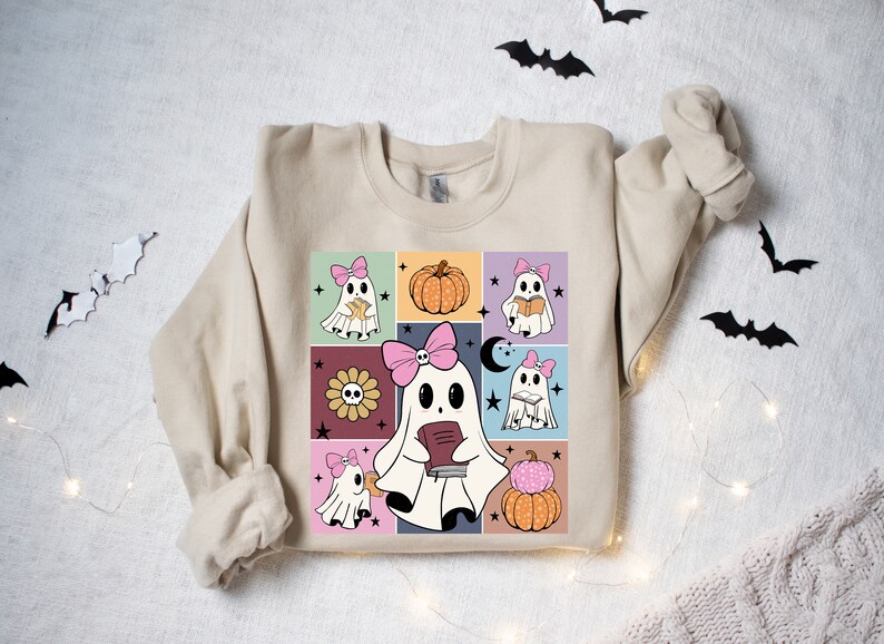 May include: Beige sweatshirt featuring a colorful graphic of cartoon ghosts with pink bows, pumpkins, and flowers. The design is set against a grid of squares with a retro aesthetic, perfect for Halloween.