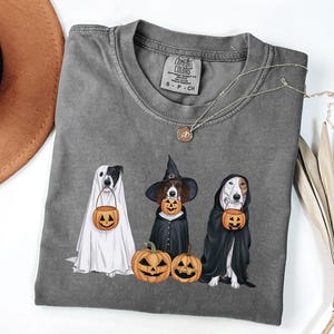 May include: A grey t-shirt featuring a graphic of three dogs dressed up for Halloween. The dogs are wearing a ghost costume, a witch costume, and a vampire costume. Each dog is holding a pumpkin bucket. The text "COMFORT COLORS" is on the tag.