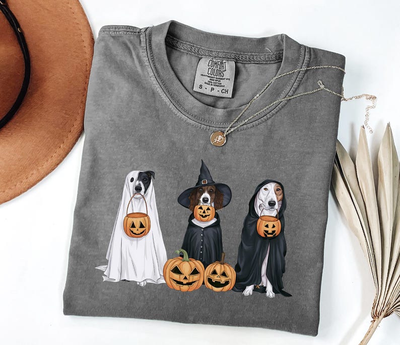 May include: A grey t-shirt featuring a graphic of three dogs dressed up for Halloween. The dogs are wearing a ghost costume, a witch costume, and a vampire costume. Each dog is holding a pumpkin bucket. The text "COMFORT COLORS" is on the tag.