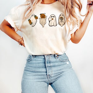 May include: A white t-shirt featuring a graphic of four ghosts. The ghosts are wearing various accessories, including a pumpkin, a flower, and a bouquet of flowers. All the ghosts are smiling.