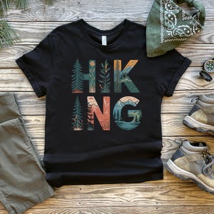 May include: Black t-shirt with a graphic print of the word "HIKING" in colorful letters with nature scenes. The letters are surrounded by trees, mountains, and a sunset.
