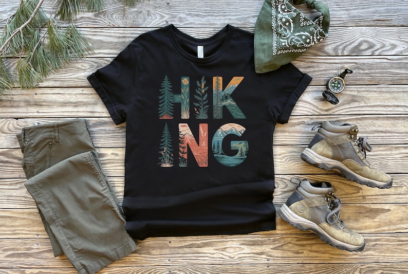 May include: Black t-shirt with a graphic print of the word "HIKING" in colorful letters with nature scenes. The letters are surrounded by trees, mountains, and a sunset.