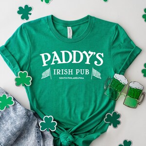 May include: Green t-shirt with white text that reads "Paddy's Irish Pub South Philadelphia". The shirt is laying on a white surface with green shamrock cutouts.