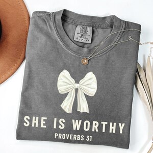May include: Gray t-shirt with a white bow and the text "SHE IS WORTHY PROVERBS 31" printed on the front.