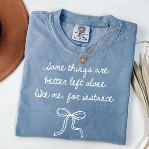May include: A blue Comfort Colors t-shirt with the text "Some things are better left alone like me, for instance" in white script. A gold necklace with a pendant rests on the shirt. A brown hat is visible in the background.