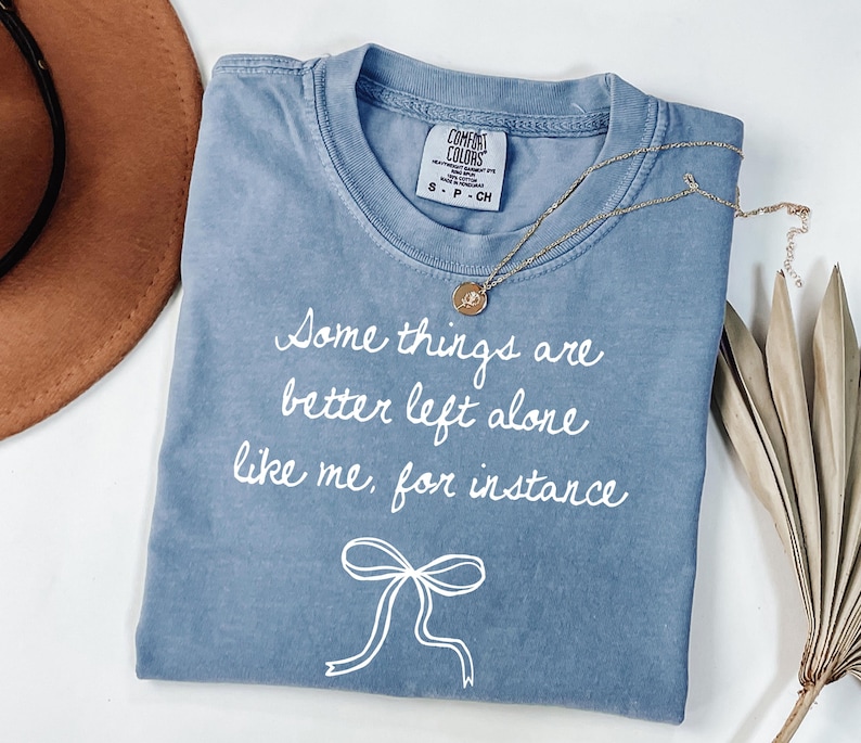 May include: A blue Comfort Colors t-shirt with the text "Some things are better left alone like me, for instance" in white script. A gold necklace with a pendant rests on the shirt. A brown hat is visible in the background.