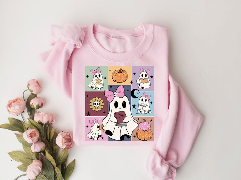 May include: A light pink sweatshirt featuring a colorful graphic of cartoon ghosts with pink bows, pumpkins, and a sunflower. The ghosts are reading books. The design is set against a grid of various colors.