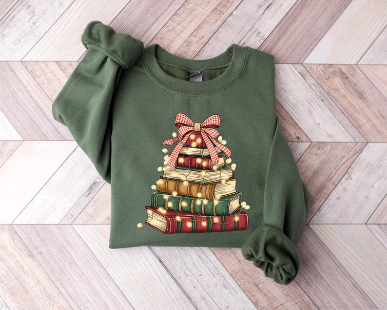 May include: Green sweatshirt featuring a stack of books designed as a Christmas tree. The books are red, brown, and green, adorned with a plaid bow and string lights. The sweatshirt has long sleeves and a crew neck.