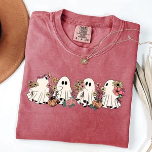 May include: A red-violet Comfort Colors t-shirt featuring a graphic of four cartoon ghosts with flowers and pumpkins. The ghosts have black eyes and are surrounded by colorful flowers. A gold necklace with a pendant is on the shirt.
