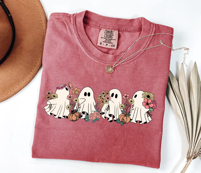 May include: A red-violet Comfort Colors t-shirt featuring a graphic of four cartoon ghosts with flowers and pumpkins. The ghosts have black eyes and are surrounded by colorful flowers. A gold necklace with a pendant is on the shirt.
