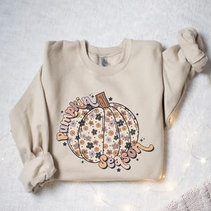K&ouml;nnte beinhalten: Ein beiger Pullover mit einem floralen K&uuml;rbisdesign und dem Text "Pumpkin Season" in einer Retro-Schrift.