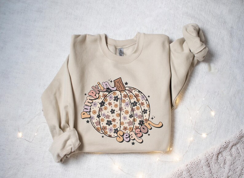 K&ouml;nnte beinhalten: Ein beiger Pullover mit einem floralen K&uuml;rbisdesign und dem Text "Pumpkin Season" in einer Retro-Schrift.