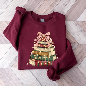 May include: Burgundy sweatshirt featuring a stack of books arranged in a Christmas tree shape. The books are adorned with string lights and topped with a red and white plaid bow. The sweatshirt has a crew neck and long sleeves.