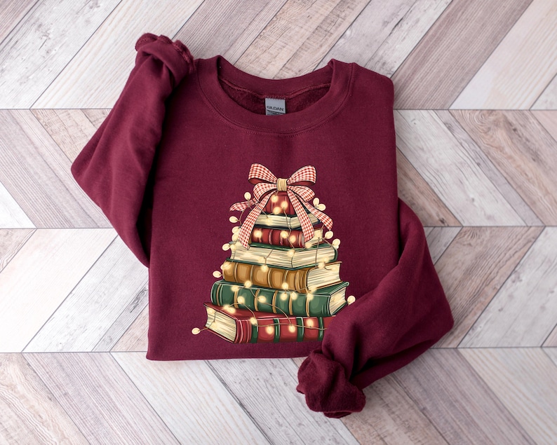 May include: Burgundy sweatshirt featuring a stack of books arranged in a Christmas tree shape. The books are adorned with string lights and topped with a red and white plaid bow. The sweatshirt has a crew neck and long sleeves.