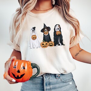 May include: A white t-shirt featuring a design of three dogs dressed in Halloween costumes. The dogs are wearing a ghost costume, a witch costume, and a vampire costume. The dogs are holding pumpkins. The t-shirt is being worn by a person with long blonde hair.