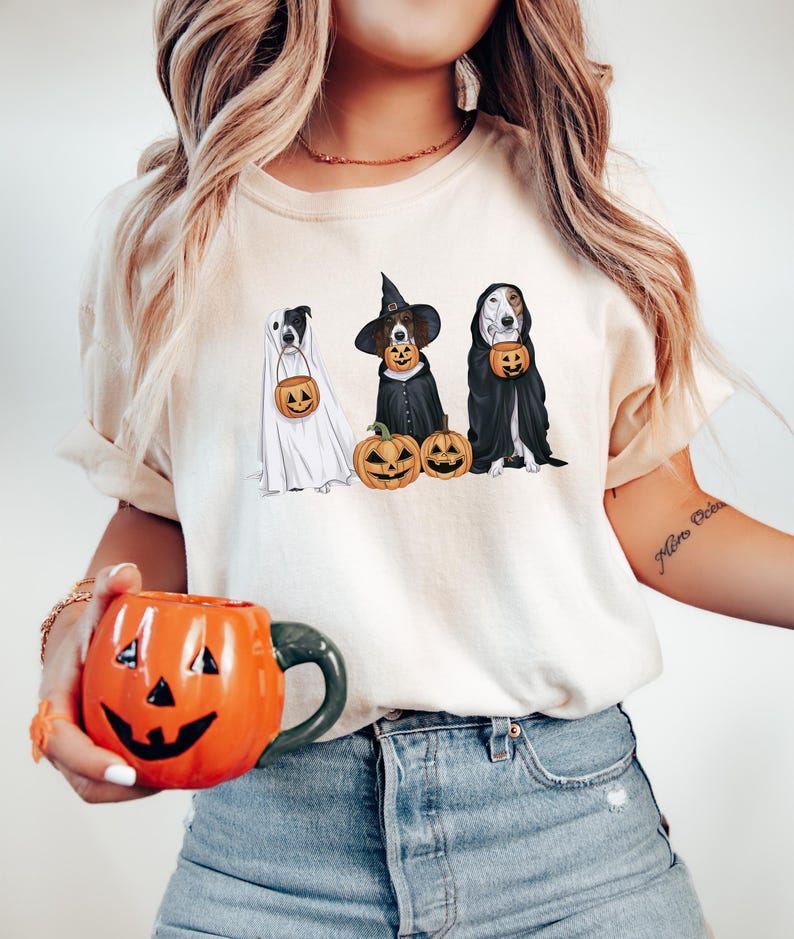 May include: A white t-shirt featuring a design of three dogs dressed in Halloween costumes. The dogs are wearing a ghost costume, a witch costume, and a vampire costume. The dogs are holding pumpkins. The t-shirt is being worn by a person with long blonde hair.