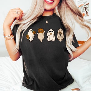 May include: A black t-shirt featuring a graphic of four ghosts. One ghost is wearing a pumpkin, one is holding a camera, and one is holding flowers. The ghosts are white with black outlines.