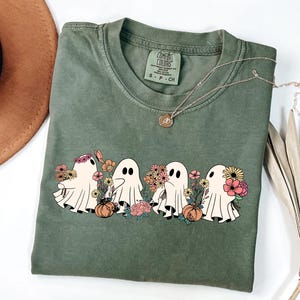 May include: Olive green t-shirt featuring a graphic of four cartoon ghosts holding flowers and pumpkins. The ghosts are in a row, with a variety of colorful flowers. A gold necklace and a brown hat are also in the image.