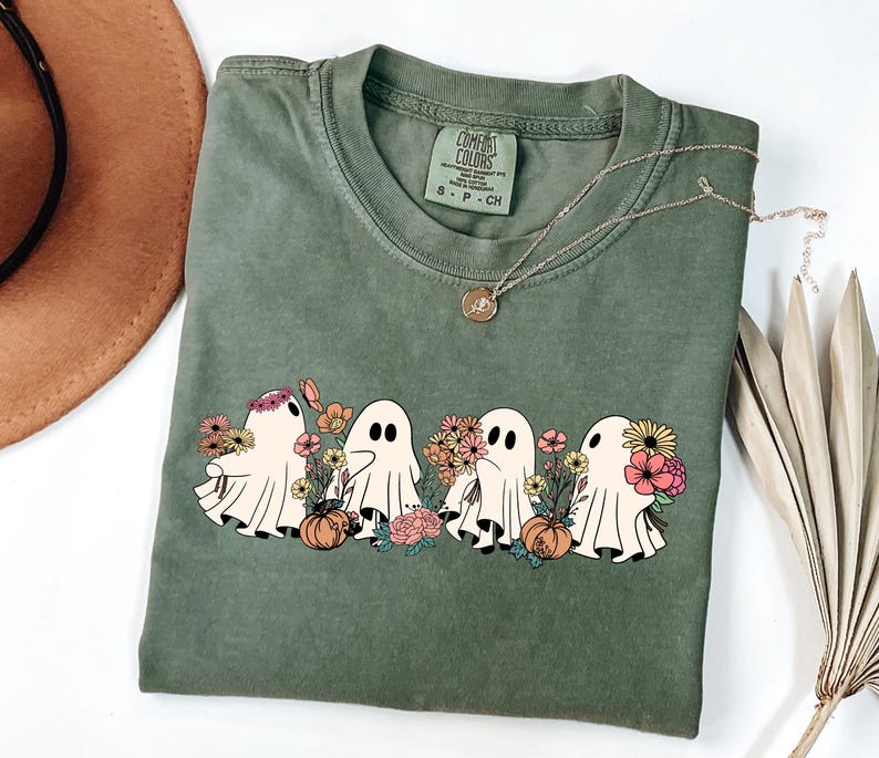 May include: Olive green t-shirt featuring a graphic of four cartoon ghosts holding flowers and pumpkins. The ghosts are in a row, with a variety of colorful flowers. A gold necklace and a brown hat are also in the image.