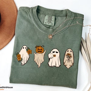 May include: A green t-shirt featuring a graphic of four ghosts. One ghost is holding a pumpkin, another is holding flowers, and the other two are smiling. The text "Comfort Colors" is visible on the tag.