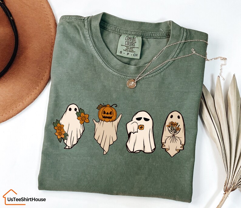 May include: A green t-shirt featuring a graphic of four ghosts. One ghost is holding a pumpkin, another is holding flowers, and the other two are smiling. The text "Comfort Colors" is visible on the tag.