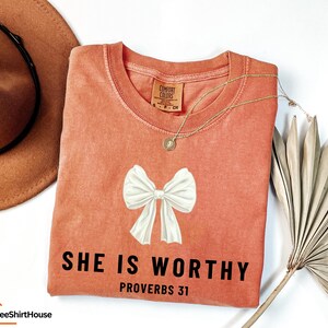 May include: A peach-colored t-shirt with a white bow and the text "SHE IS WORTHY PROVERBS 31" in black.