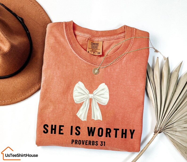 May include: A peach-colored t-shirt with a white bow and the text "SHE IS WORTHY PROVERBS 31" in black.