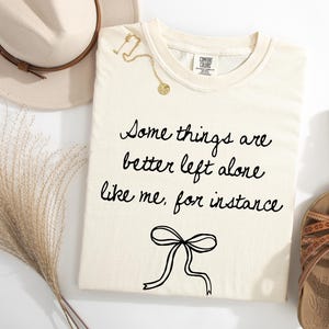 May include: Cream-colored t-shirt with the phrase "Some things are better left alone like me, for instance" in black script. A gold necklace and brown sandals are also visible. A beige hat and dried grass are in the background.