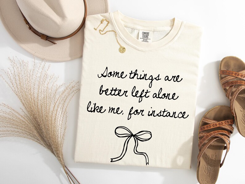 May include: Cream-colored t-shirt with the phrase "Some things are better left alone like me, for instance" in black script. A gold necklace and brown sandals are also visible. A beige hat and dried grass are in the background.