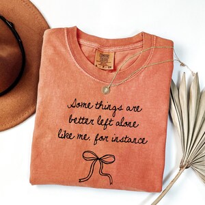 May include: Coral-colored t-shirt with the text "Some things are better left alone like me, for instance" in black script. A gold necklace with a pendant and a brown hat are next to the shirt. A dried palm leaf is on the right.