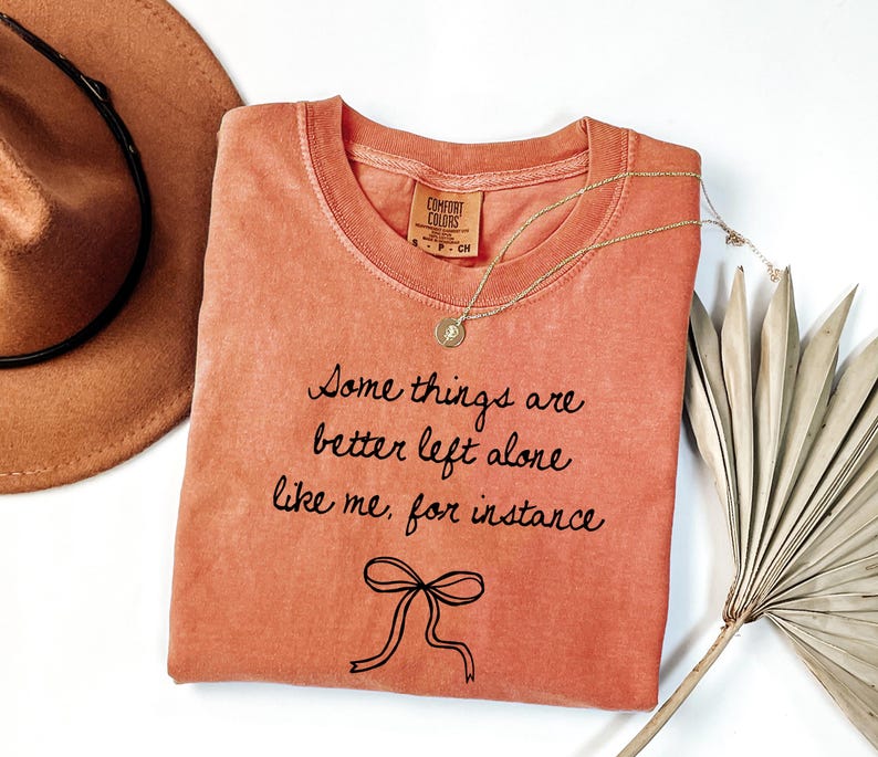 May include: Coral-colored t-shirt with the text "Some things are better left alone like me, for instance" in black script. A gold necklace with a pendant and a brown hat are next to the shirt. A dried palm leaf is on the right.