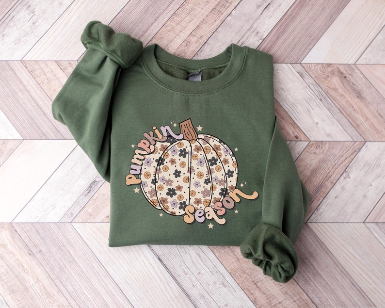 K&ouml;nnte beinhalten: Ein gr&uuml;nes Sweatshirt mit einem floralen K&uuml;rbisdesign und dem Text "pumpkin season" in einer Retro-Schrift.
